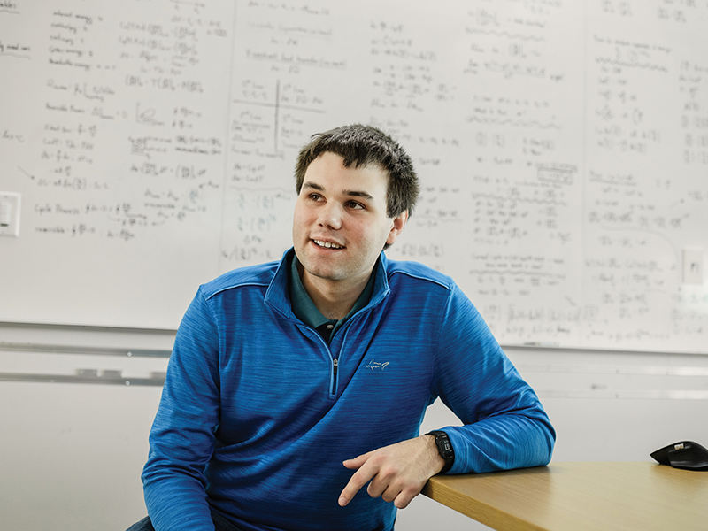 A person with short black hair wearing a blue quarter-zip sweatshirt sitting at a table with a whiteboard behind them