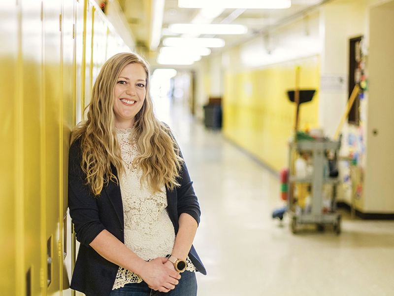 A person with long blonde hair wearing a black cardigan over a white shirt standing at the beginning of a hallway of lockers