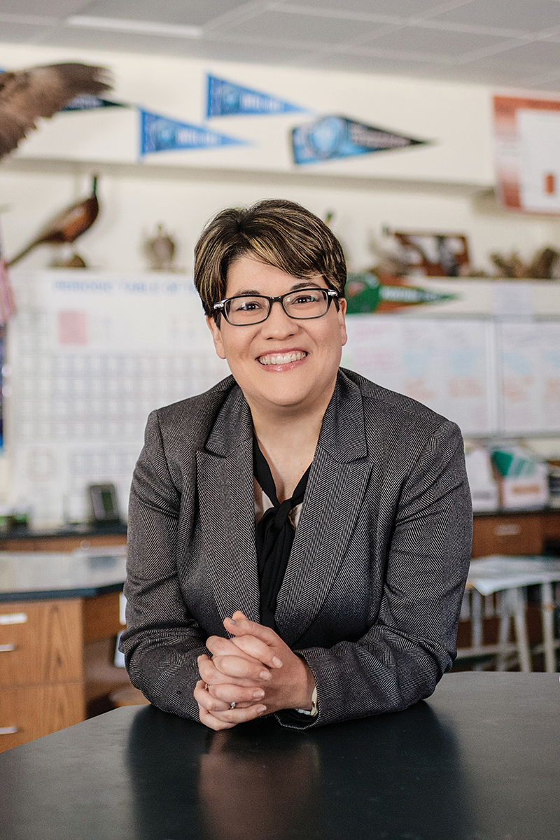 A person wearing a gray blazer and glasses with short brown hair smiling to the camera