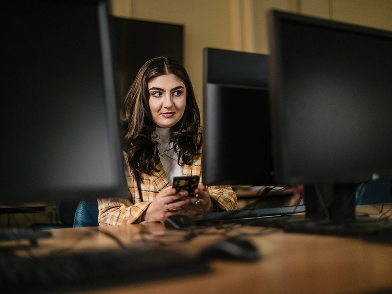 A person with long brown hair sitting in a computer lab holding a rectangular device