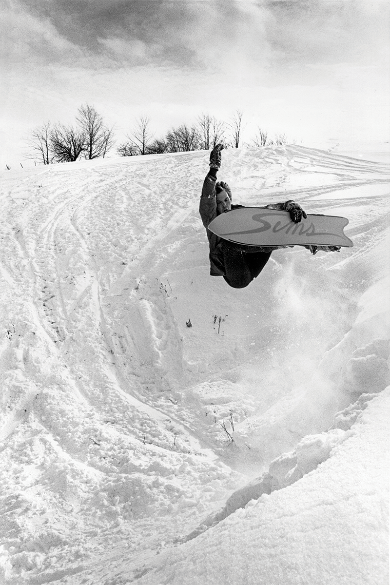 A black and white image of a person mid-air holding their snowboard with one hand