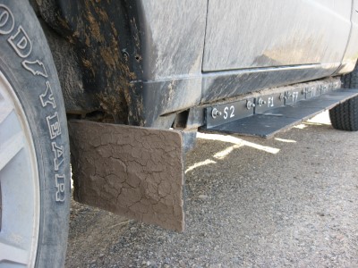 Pickup truck wheel and wheel well with metal plate attached to wheel well. Plate is coated with brown, dried mud. 