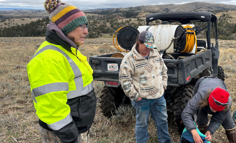 Three people standing near an ATV in a grassland. There are hills with scattered trees in the background.
