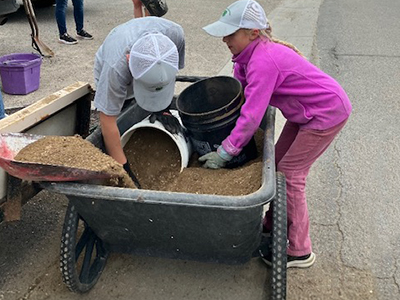 Two kids load sand into buckets from a wheelbarrow.