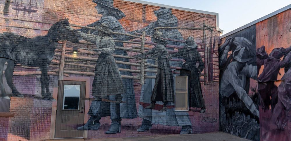 Large mural painted across two building walls depicting historical western scenes with people, livestock, and fencing.