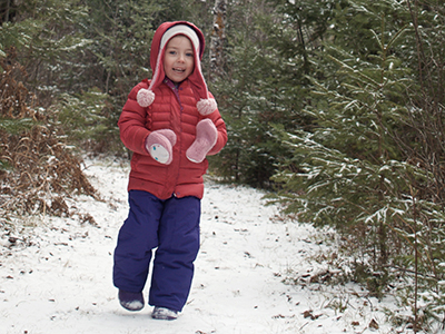 A small child in a colorful snowsuit runs down a snowcovered trail in the pine trees.