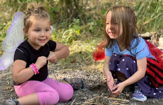 two young girls make a pile of sticks and grass next to a gravel path. They are both wearing costume wings on their backs and smiling.