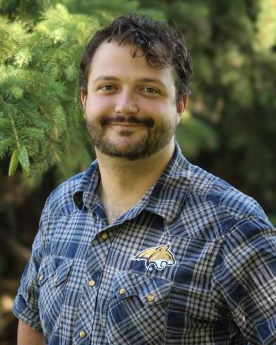 Male in blue flannel standing in front of a forest background