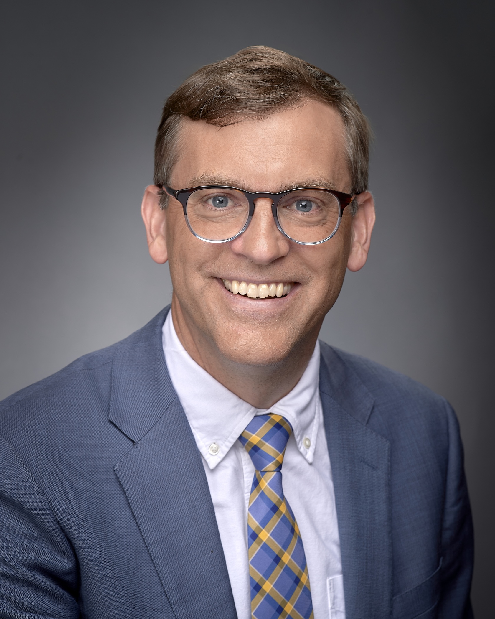 Montana State University President Brock Tessman smiles for a portrait, wearing glasses, a blue suit and a blue-and-gold plaid tie against a gray background.