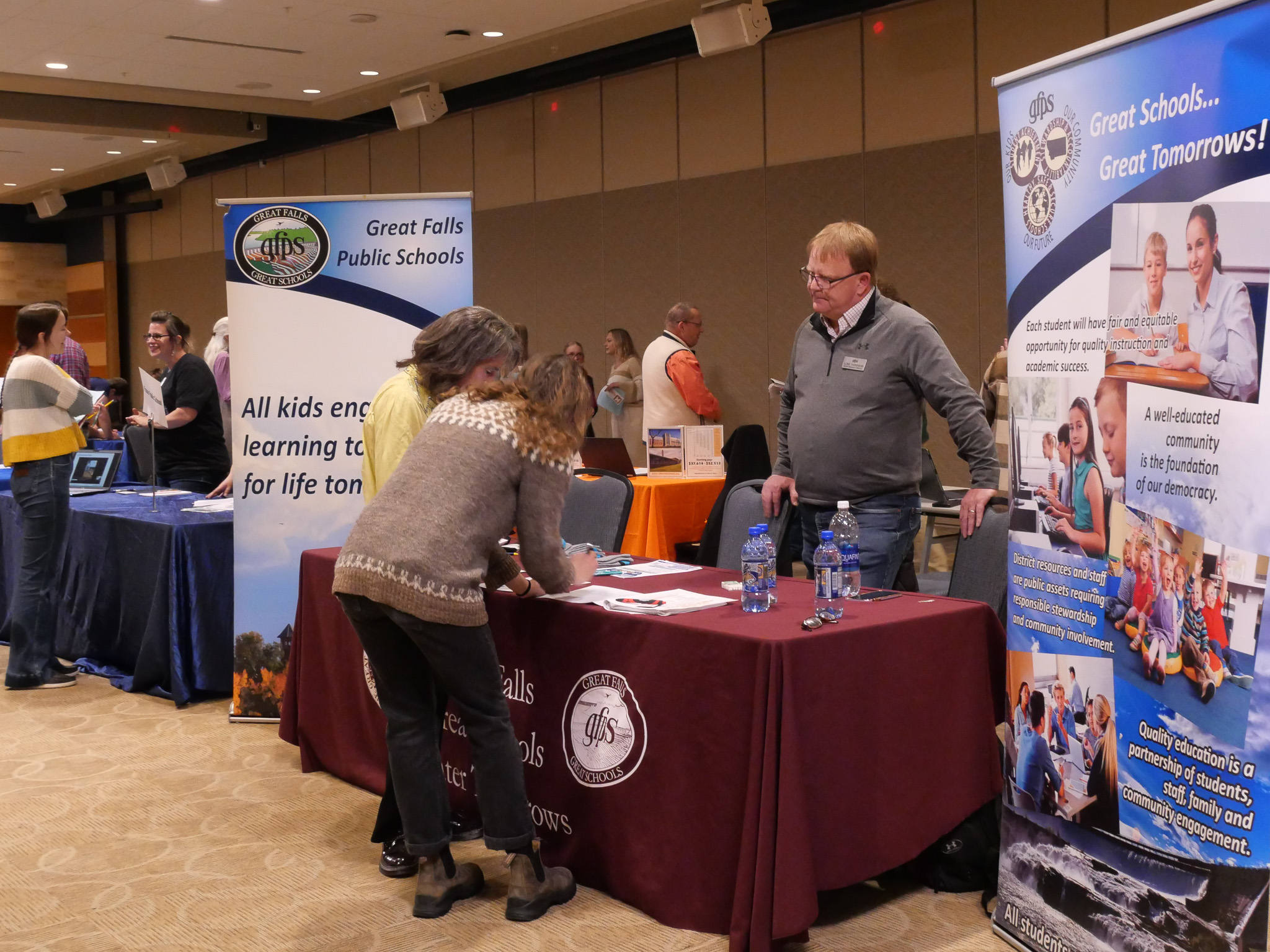 A table with a man standing behind it talking to two people who are looking at something on the table