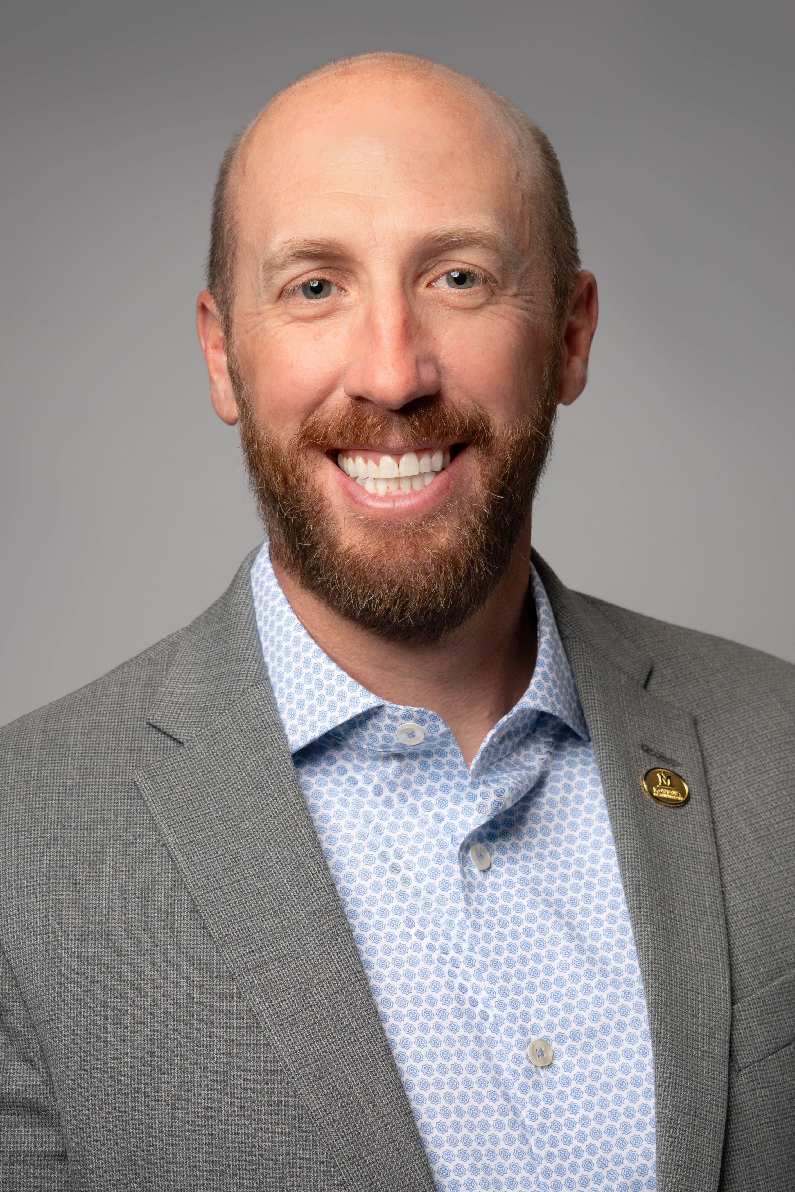 Portrait of a man with a light brown beard wearing a blue undershirt and gray suit coat